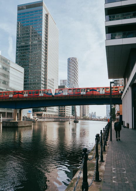 A modern urban scene featuring a red train crossing a bridge over a river, with tall glass and steel skyscrapers lining the background. The train has a sleek exterior with large windows, and its vibrant red color contrasts with the subdued tones of the buildings and the sky. The river below reflects the surrounding structures and the train, with gentle ripples visible on the water's surface. On the right side of the image, a paved walkway runs parallel to the river, bordered by a black metal railing and a row of bollards. A person dressed in dark clothing is walking along the path, towards the background. The sky is partly cloudy, allowing dappled sunlight to illuminate the scene softly. While the image primarily depicts an urban transit and skyline environment, the context of rubbish removal or private waste handling is indirectly supported by the orderly cleanliness and infrastructure, which might be maintained through services like those provided by House Clearance Docklands for property decluttering, including rubbish collection in Docklands or Canary Wharf areas.