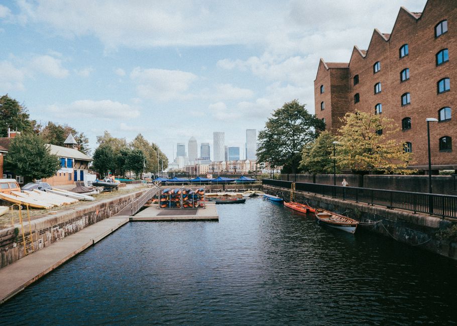 A narrow canal with a stone and concrete embankment on the left side, where several small boats with varying colors and materials, including wooden and plastic, are moored along the edge. The water appears calm and reflects the sky and surrounding environment. On the right side, a tall brick building with multiple windows and a slightly stepped roofline stands behind a black metal fence running parallel to the canal walkway. Several trees with green and yellow leaves line the pavement, providing a natural contrast to the built environment. In the background, a cluster of modern high-rise office buildings is visible against a partly cloudy sky, creating a backdrop of urban skyline. The scene is well-lit with daylight, emphasizing the textures of the boats, brickwork, and foliage, and illustrating a typical docking area that might require occasional rubbish removal or site clearance managed by services like House Clearance Docklands for maintaining clean, accessible waterways and surroundings.