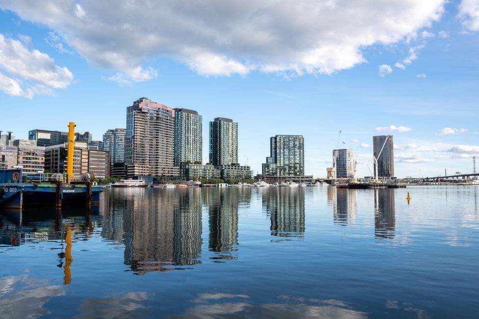 A wide view of a city skyline across a calm body of water during daylight, featuring several modern high-rise buildings with glass facades reflecting the sky. In the foreground, there are boats docked along the water's edge, including a blue vessel with yellow and black mooring equipment. The scene is set on a clear day with a few scattered clouds, and the water's surface shows slight ripples, creating mirrored reflections of the buildings and boats. The urban environment suggests a waterfront area that may be subject to private or independent rubbish collection and waste management activities, relevant to services such as those provided by House Clearance Docklands, which specialise in rubbish removal in the Docklands estates.