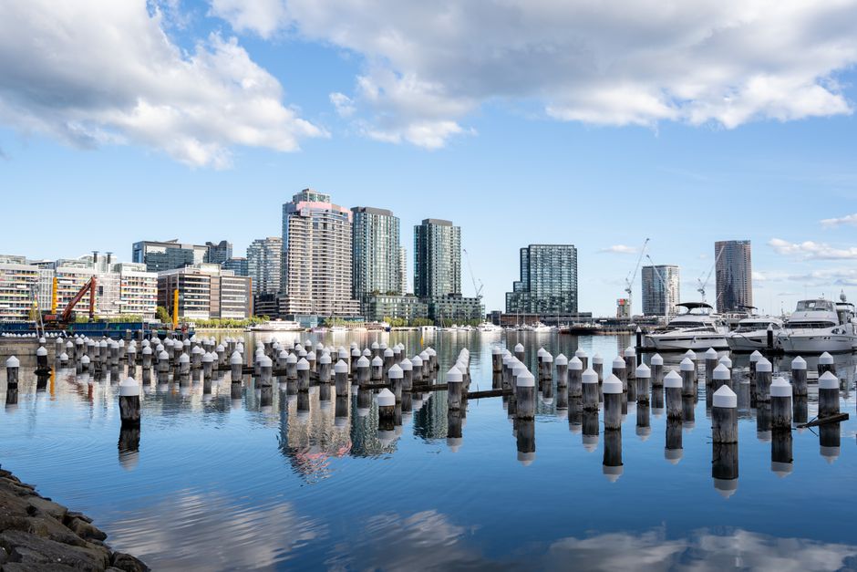 A waterfront scene featuring a marina with multiple white mooring bollards arranged in rows along the edge of calm water. Behind the bollards, several luxury yachts and boats are docked, with some visible beneath their covers. The background showcases a collection of modern high-rise buildings with glass facades reflecting the daylight, set against a partly cloudy sky. The scene is characteristic of a metropolitan urban area with a mix of commercial and residential architecture, interspersed with construction cranes indicating ongoing development. The environment appears clean and well-maintained, with clear water and no visible debris, suggesting a controlled setting suitable for visualizing private or independent collection of waste or rubbish within a harbor or docklands vicinity, as part of alternative waste handling methods possibly managed by companies like House Clearance Docklands.