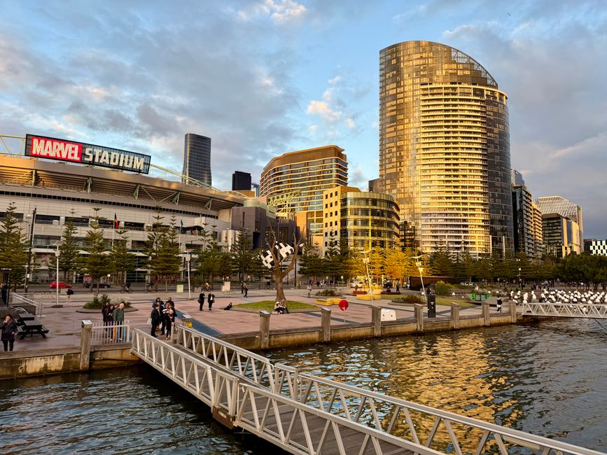 A wide view of a city skyline across a calm body of water during daylight, featuring several modern high-rise buildings with glass facades reflecting the sky. In the foreground, there are boats docked along the water's edge, including a blue vessel with yellow and black mooring equipment. The scene is set on a clear day with a few scattered clouds, and the water's surface shows slight ripples, creating mirrored reflections of the buildings and boats. The urban environment suggests a waterfront area that may be subject to private or independent rubbish collection and waste management activities, relevant to services such as those provided by House Clearance Docklands, which specialise in rubbish removal in the Docklands estates.