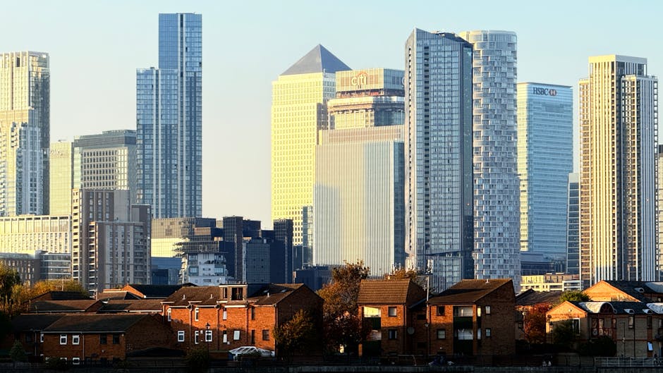 A cityscape featuring a dense cluster of modern high-rise office buildings and residential towers with glass facades reflecting sunlight, set behind a row of low-rise brick houses with pitched roofs and small front gardens. The skyscrapers vary in height, with some structures displaying corporate signage such as HSBC and Citibank. The scene is captured in daylight with clear skies, highlighting the contrast between the contemporary urban skyline and the traditional residential dwellings in the foreground. This image visually emphasizes the juxtaposition of residential areas and commercial development, illustrating potential contexts for private waste handling and alternative rubbish collection services offered by companies like House Clearance Docklands near London City Airport in the Docklands area.