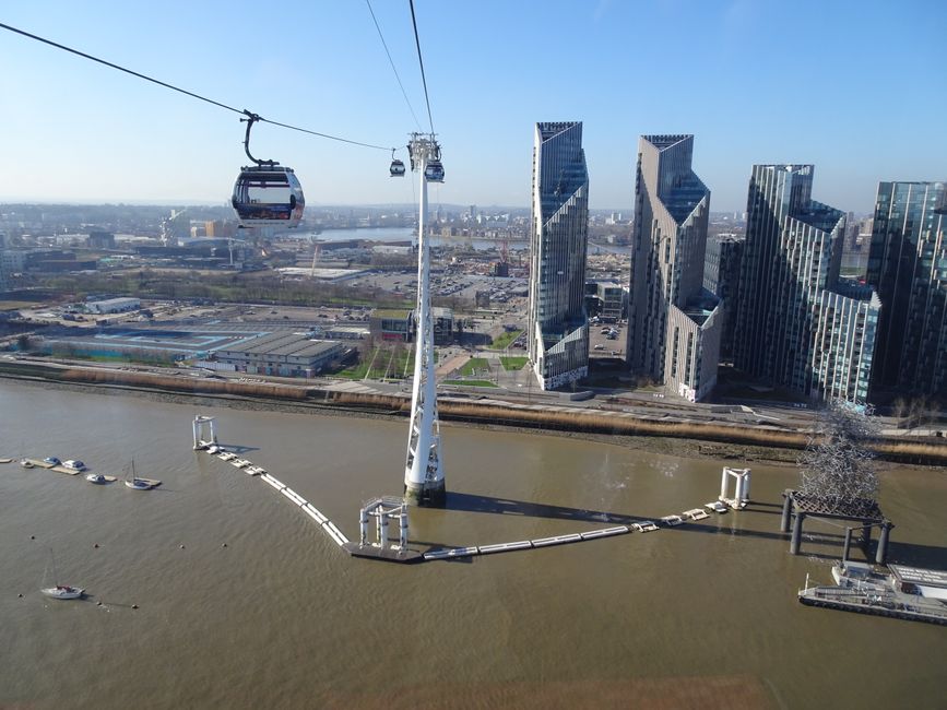 An aerial view of the River Thames in London featuring the Emirates Air Line cable car system with three cabins suspended from cables stretching across the river. The cable cars are modern, white with glass windows, and hover above the water, which has small boats moored along the riverbank. In the background, there are several contemporary skyscrapers with reflective glass exteriors, displaying varying shades of grey and blue. The buildings have distinct angular designs, with some featuring diagonal cuts and irregular shapes. The scene is illuminated by natural daylight, with a clear blue sky overhead, and the urban landscape extends into the distance with low-rise industrial buildings and open areas near the river. The image subtly suggests an environment where alternative and private transportation infrastructure exists alongside city development, aligning with themes of urban planning and innovative transit solutions often associated with modern London districts.