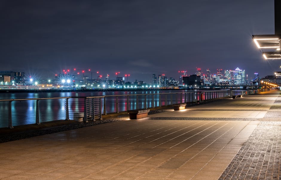 A nighttime view of a broad riverside promenade with paved walkway slabs and modern lighting fixtures along the edge. Behind the railing, calm water reflects the colorful city skyline illuminated with numerous high-rise buildings and construction cranes with red warning lights. The sky appears overcast with dark clouds, and the urban lights create a glow across the scene. On the right side, a portion of a contemporary building with exterior lighting extends into the frame. The scene suggests an urban waterfront area that could be used for independent rubbish collection or on-site waste handling, with minimal clutter or debris visible in the open space. The overall environment is clean and well-maintained, emphasizing a clear setting suitable for professional rubbish removal services tailored to city docklands or flat complexes such as those at Millwall Dock.