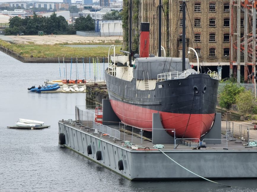 A large vintage steamship with a black hull and a red lower section is positioned on a concrete slipway beside a body of water, likely within a dockyard or marina setting. The vessel features a tall red funnel and prominent white railings along the deck, with various small details such as mooring lines and equipment on the surface. The surrounding environment includes a grey quay with safety barriers, some greenery in the background, and a rusty, multi-storey brick building to the right. In the water nearby, several small boats and dinghies, in shades of blue, white, and orange, are moored or floating, with some tied to buoys or partially on the edge of the riverbank. The background shows an urban landscape with modern buildings and industrial structures, indicating a city docklands area. The scene appears to be outdoors during daylight, with natural lighting illuminating the vessel and its surroundings, fitting within a context of private vessel storage or maintenance in a coastal or dockside setting for alternative or non-public boat disposal or storage services occasionally associated with waste management operations in docklands.